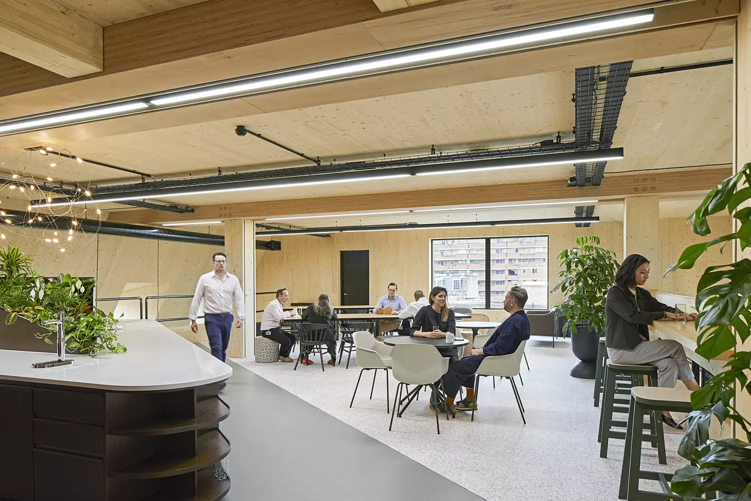 Modern office environment featuring an open-plan layout with multiple people seated at tables. The space includes large windows that bring in natural light, exposed ceiling beams with industrial-style lighting and a mix of functional furniture such as chairs, tables and a counter with shelving. Potted plants are placed throughout the room, adding a natural touch to the contemporary and collaborative workspace design.