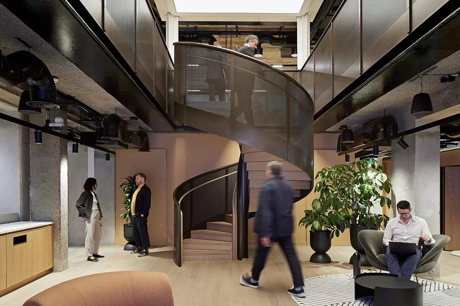 Modern office interior with a central spiral staircase as a key architectural feature. The space includes contemporary furniture such as chairs and tables, along with a potted plant adding a touch of greenery. The open-plan layout is enhanced by natural light streaming in from above, and the overall design showcases clean lines and a minimalist aesthetic that promotes openness and movement.