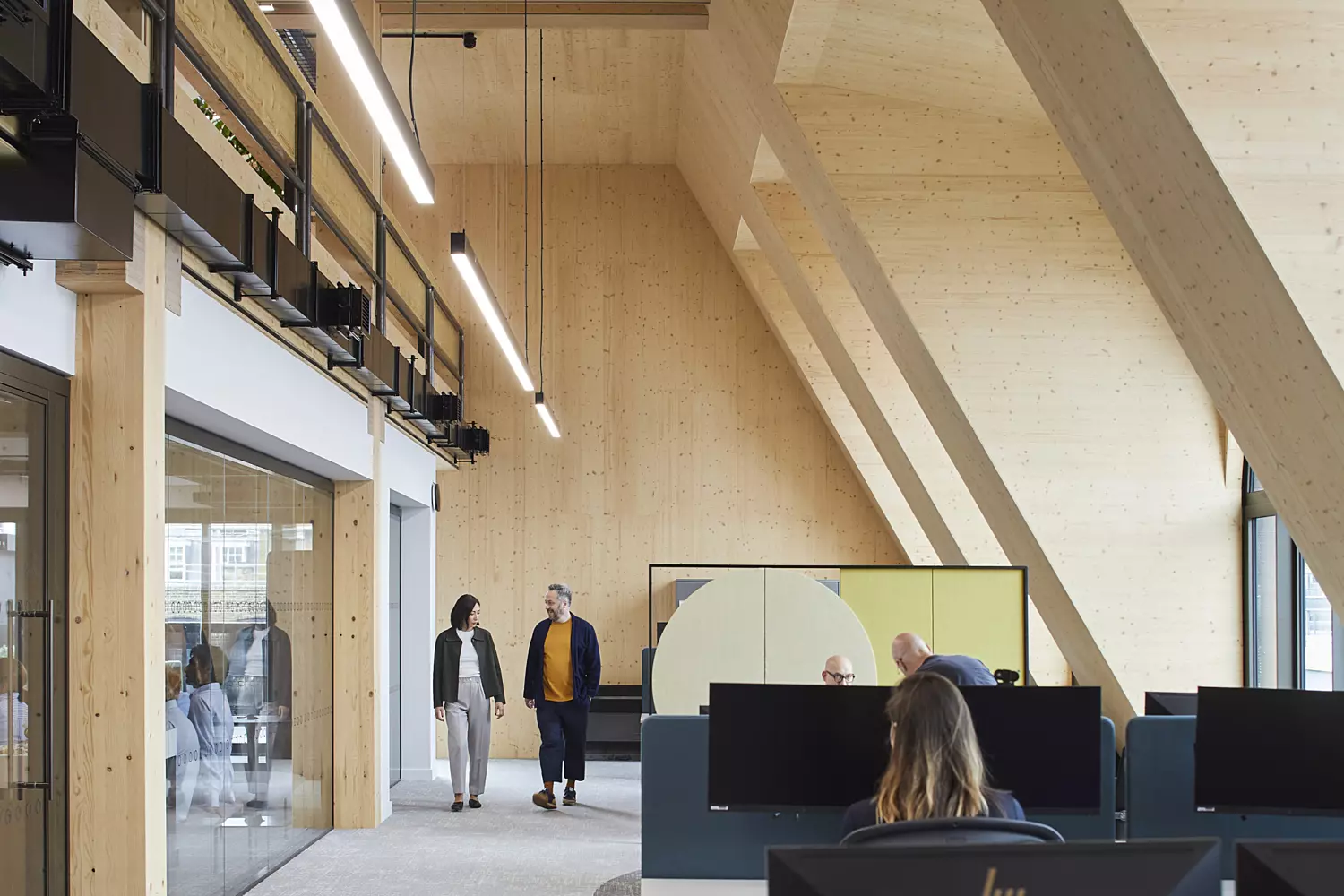 Modern office interior featuring high wooden ceilings and expansive windows that allow natural light to fill the space. The design is clean and minimalist, incorporating light-coloured wood and glass elements. Several people are present, either seated at desks with computer monitors or standing, contributing to a dynamic yet uncluttered working environment.