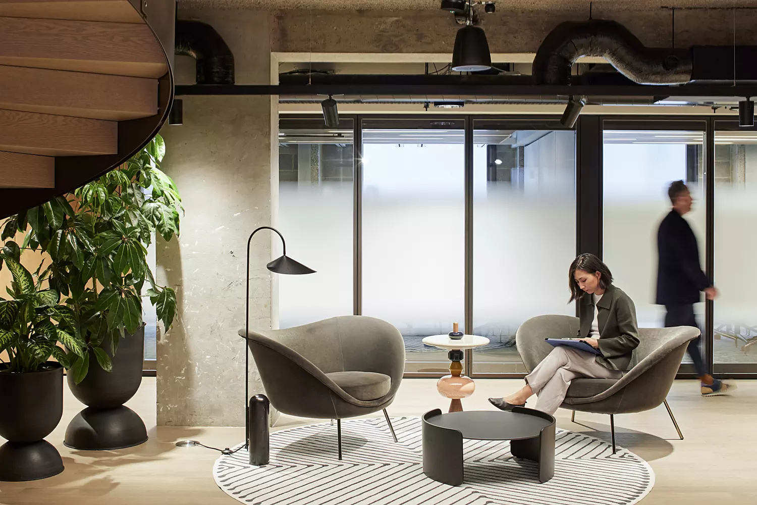 Modern office lounge area featuring two grey armchairs and a small round table placed on a circular rug. The space includes large frosted-glass windows that diffuse natural light, potted plants for a touch of greenery, and an industrial-style ceiling with exposed pipes. The design combines comfort with a contemporary, minimalist aesthetic, creating a relaxed yet professional atmosphere.