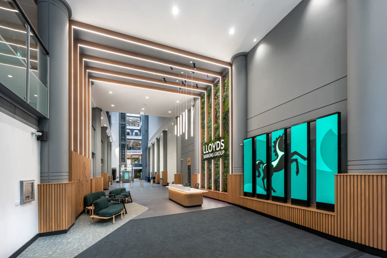 A spacious modern lobby with high ceilings and tall grey walls featuring vertical wooden slats. A series of suspended linear lights hangs above a central reception desk made of light wood. To the right, a green living wall surrounds the Lloyds Banking Group logo. Next to it, large digital screens display the company’s black horse symbol on a teal background. Soft seating with dark green upholstery sits to the left, and the far end of the space opens into a bright atrium with additional architectural details.