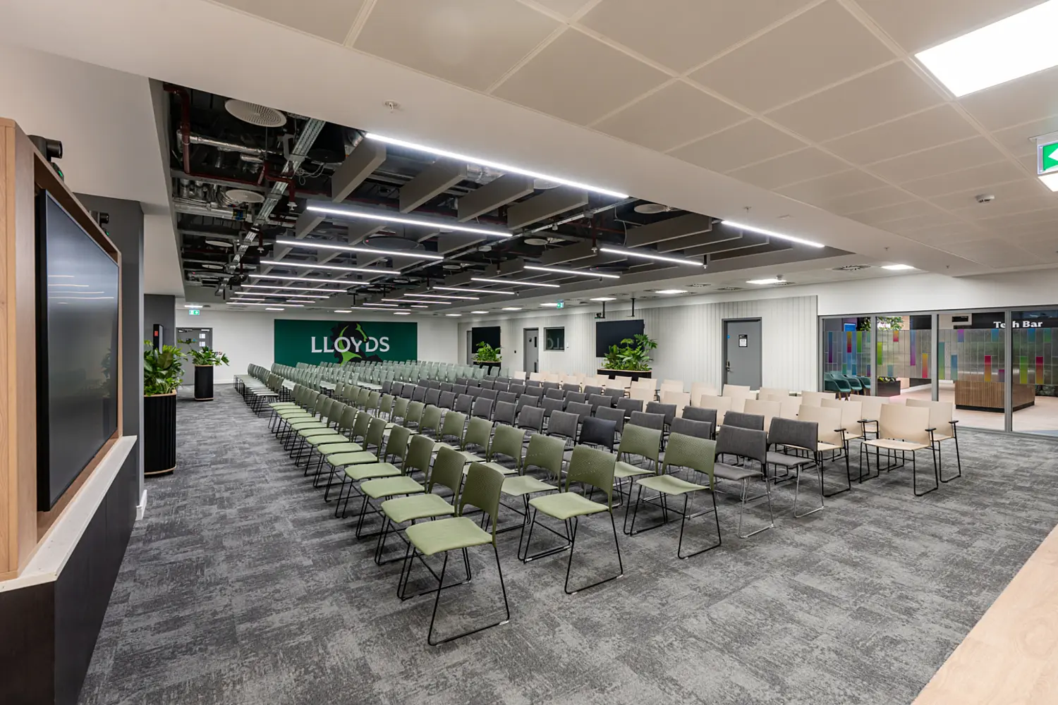 A large presentation and event space with rows of chairs arranged in a theatre layout, featuring alternating green, grey and cream seating. The room has a modern industrial ceiling with exposed services and linear LED lighting. At the front, a dark green wall displays the Lloyds name in white. The floor is covered in grey patterned carpet and there are potted plants placed around the space. To the right, glass doors lead to an area with colourful graphics, while a large screen is positioned on the left side of the room.