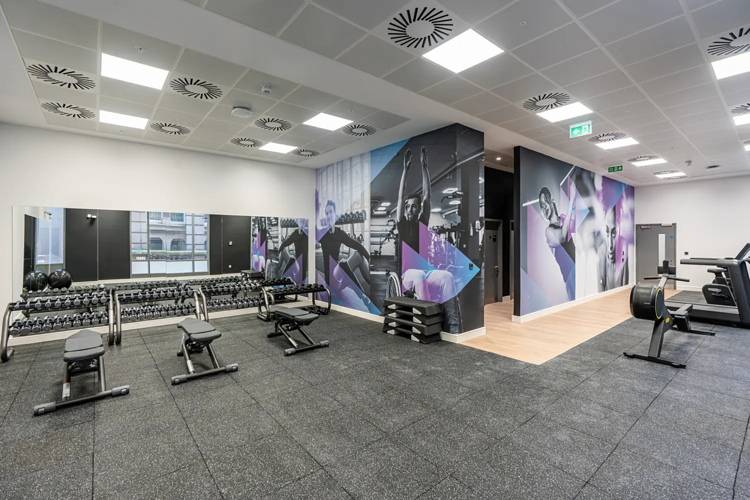 A modern corporate gym with black rubber flooring and a long wall of mirrors above a full rack of dumbbells. Adjustable weight benches are spaced across the floor. The central column and surrounding walls feature large fitness-themed graphics in shades of blue, purple and black, showing people exercising. To the right, a lighter wood floor section leads to additional gym equipment, including cardio machines and an indoor rowing machine. The ceiling has a grid of square lights and circular ventilation units, giving the space a clean and well-organised appearance.