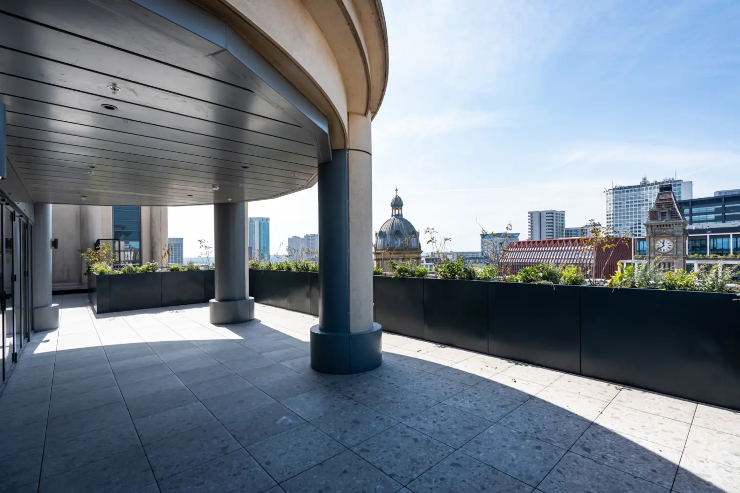 A spacious rooftop terrace with large grey stone tiles and a curved overhang supported by round columns. A long planter filled with greenery runs along the terrace edge. Beyond the terrace, there is a clear view of surrounding Birmingham city centre, including a domed historic building and a clock tower. Modern high‑rise buildings stand further in the distance under a bright blue sky.