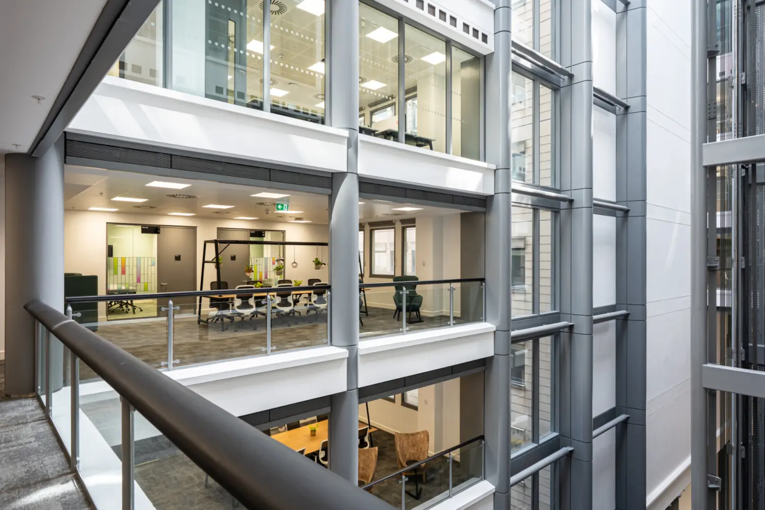 An internal atrium with multiple office floors visible through large glass panels framed by grey structural beams. Each level contains modern workspaces, including meeting rooms, desks and seating areas. The walkways have glass balustrades with dark handrails. Bright ceiling lights illuminate the interior and create reflections on the glass. Lift shafts and vertical structural supports run up the right side of the atrium. The overall design shows an open multi‑storey office environment with clear sightlines across different floors.