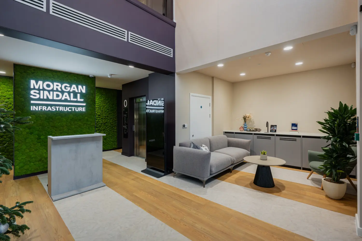 Reception area at Morgan Sindall Infrastructure featuring a moss green branded feature wall, a concrete-style reception desk and a contemporary seating area with a grey sofa, armchair and round coffee table. Light wood flooring, indoor plants and high ceilings create a welcoming, modern office entrance.