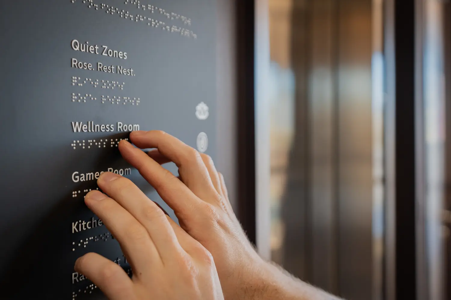 Close-up of a person reading a tactile wayfinding sign with braille and raised lettering, featuring room names including Quiet Zones, Wellness Room and Games Room. The image focuses on accessibility features within a modern office environment, with softly blurred interior spaces in the background.