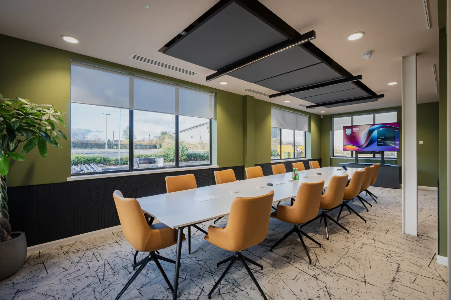 Contemporary meeting room with a long conference table surrounded by tan upholstered chairs, set against olive green walls and large windows providing natural daylight. Acoustic ceiling panels, integrated audiovisual technology and a mobile presentation screen create a modern collaborative space designed for meetings, presentations and hybrid working.