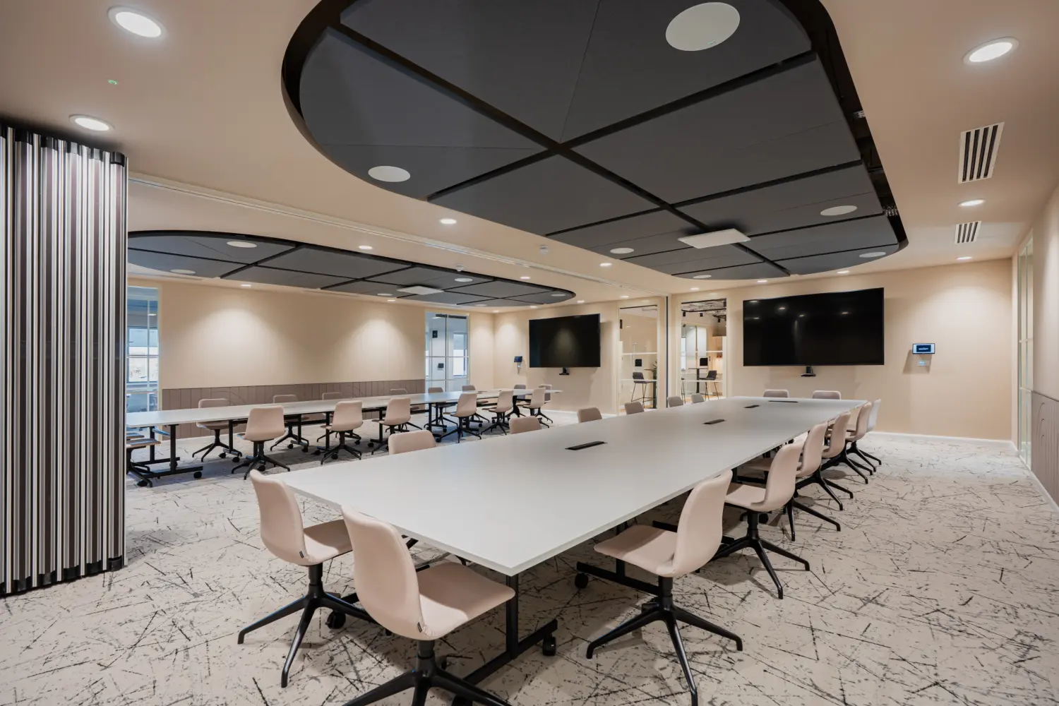 Large multi-purpose meeting and training room featuring modular white tables arranged in a U-shape and classroom layout, with ergonomic chairs, wall-mounted presentation screens and integrated video conferencing facilities. Distinctive acoustic ceiling panels, soft neutral finishes and glazed partitions create a modern, flexible workspace designed for collaboration and presentations.