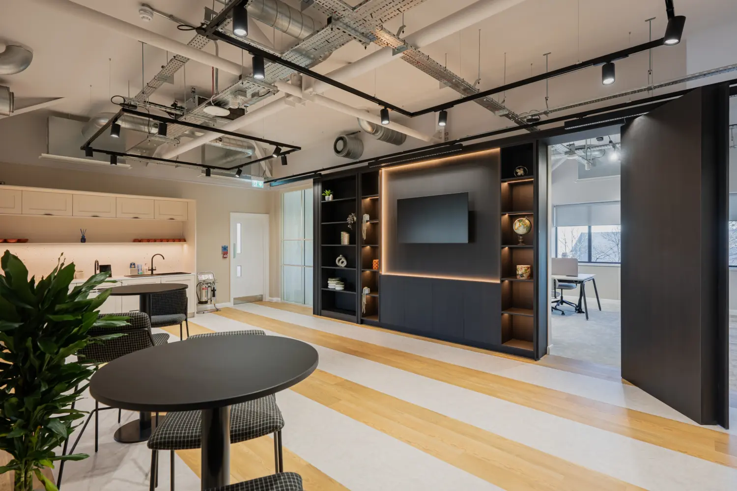 Contemporary office collaboration space featuring a kitchenette, café-style seating and a dark timber media wall with illuminated shelving, a mounted screen and a disguised doorway hidden in the shelving that reveals a secret library room. Exposed ceiling services, light wood flooring and glazed meeting rooms create an open, modern workplace environment with a mix of social and focused work areas.