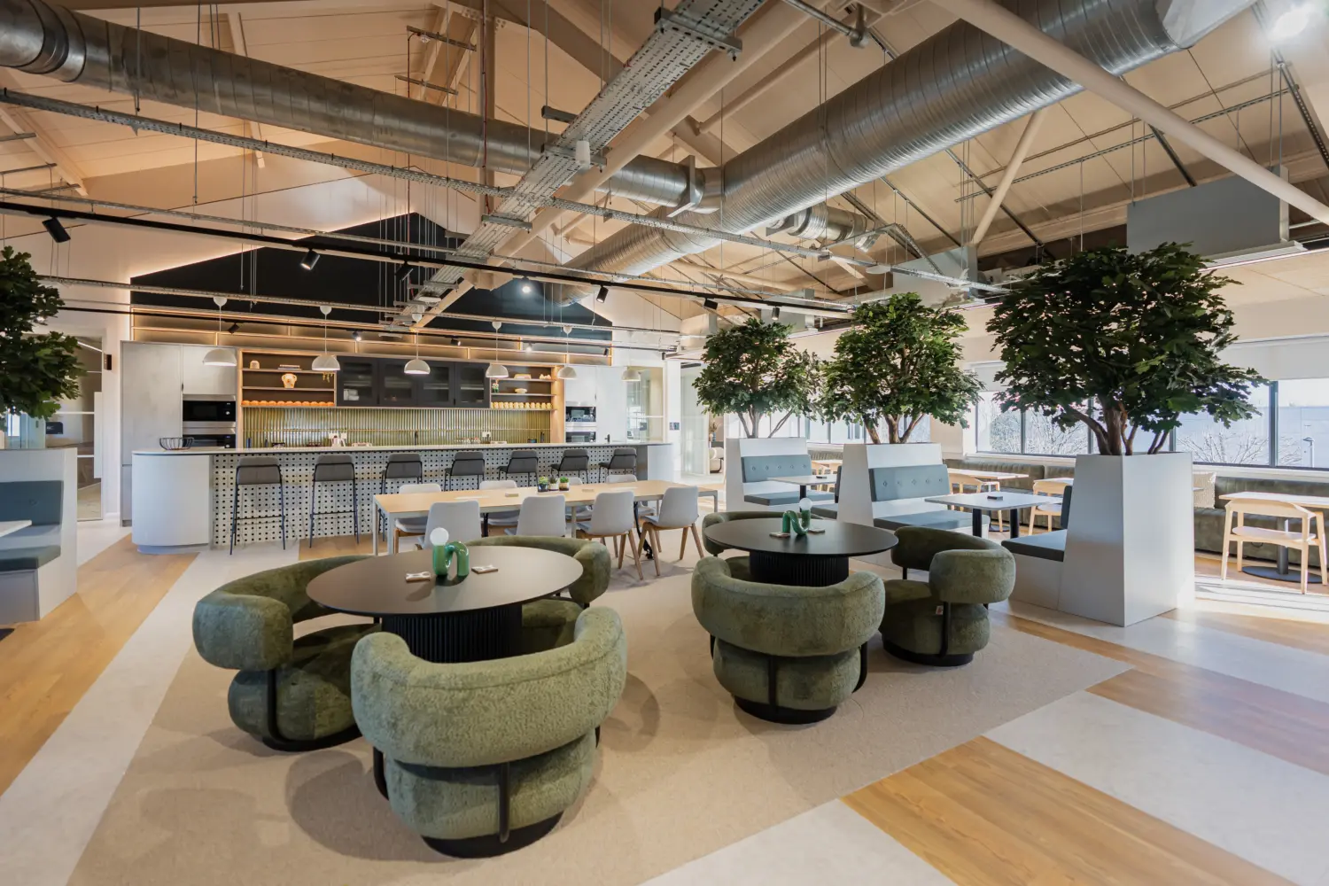 Modern open-plan office breakout and café area featuring a central kitchen island with bar seating, round tables surrounded by green upholstered lounge chairs and large indoor trees in white planters. Exposed ceiling services, light wood flooring and floor-to-ceiling windows create a bright, contemporary workspace.
