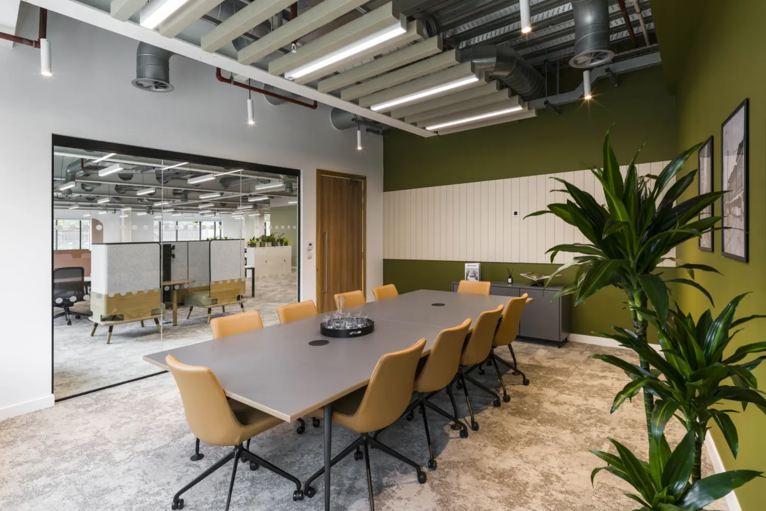 Meeting room with a long rectangular table surrounded by tan upholstered chairs on casters, set on a light grey carpet. A large potted plant stands in the foreground on the right beside a dark green accent wall with framed artwork and a white slatted acoustic panel. The ceiling features exposed services and suspended linear lighting with white baffles. A full-height glass partition on the left looks into an open office area with desks and booths, while a wooden door and low storage unit sit against the far wall. Natural light enters from windows beyond the adjacent workspace.