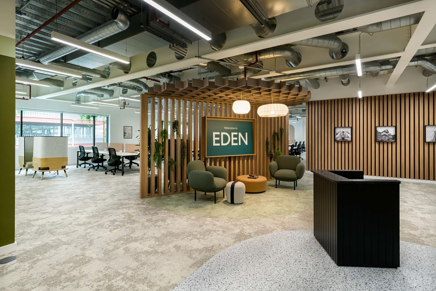 Bright, modern office reception area with an open-plan layout. In the centre, a wooden slatted feature structure frames a green sign reading “Welcome to EDEN”, with soft seating including armchairs and a round ottoman arranged beneath warm pendant lights. To the right, a dark reception desk sits against a wall of vertical wooden slats displaying framed black-and-white photos. To the left, a workspace with desks and office chairs is visible through large windows. The ceiling features exposed ductwork, pipes and linear lighting, while the floor combines carpet and polished concrete finishes, giving the space a contemporary, industrial feel.
