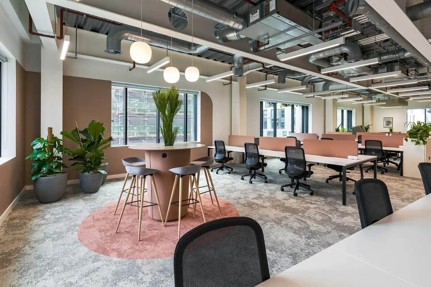 Contemporary open-plan office area with a warm, neutral palette and soft pink accents. In the foreground, a round high table with a central planter holding tall greenery sits on a circular pink rug, surrounded by high stools. To the left, additional potted plants are placed near large windows that bring in natural light and offer views of an external building.  Beyond the breakout area, rows of desks with black office chairs are arranged in clusters, divided by soft pink desk screens that add a sense of privacy while maintaining an open feel. The ceiling is exposed, featuring silver ductwork, pipes and suspended linear lighting, complemented by a row of globe pendant lights above the central table.  The flooring is a patterned carpet in light neutral tones, and the overall space combines soft furnishings, planting and industrial ceiling details to create a modern, relaxed working environment.
