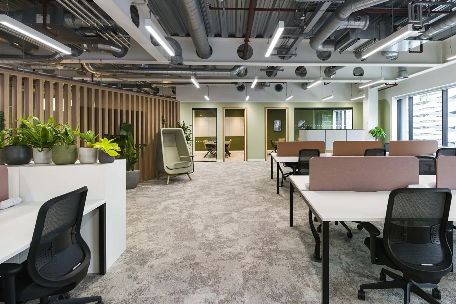 Open-plan office with rows of white desks and black mesh chairs arranged in clusters, separated by low fabric screens. A white storage unit with several green potted plants sits in the foreground on the left, next to a vertical wooden slat partition. The space features a carpeted floor, exposed ceiling with visible ductwork and linear lighting, and green-accent walls. In the background, there are glass-fronted meeting rooms and a partitioned area, with natural light coming in from large windows on the right.