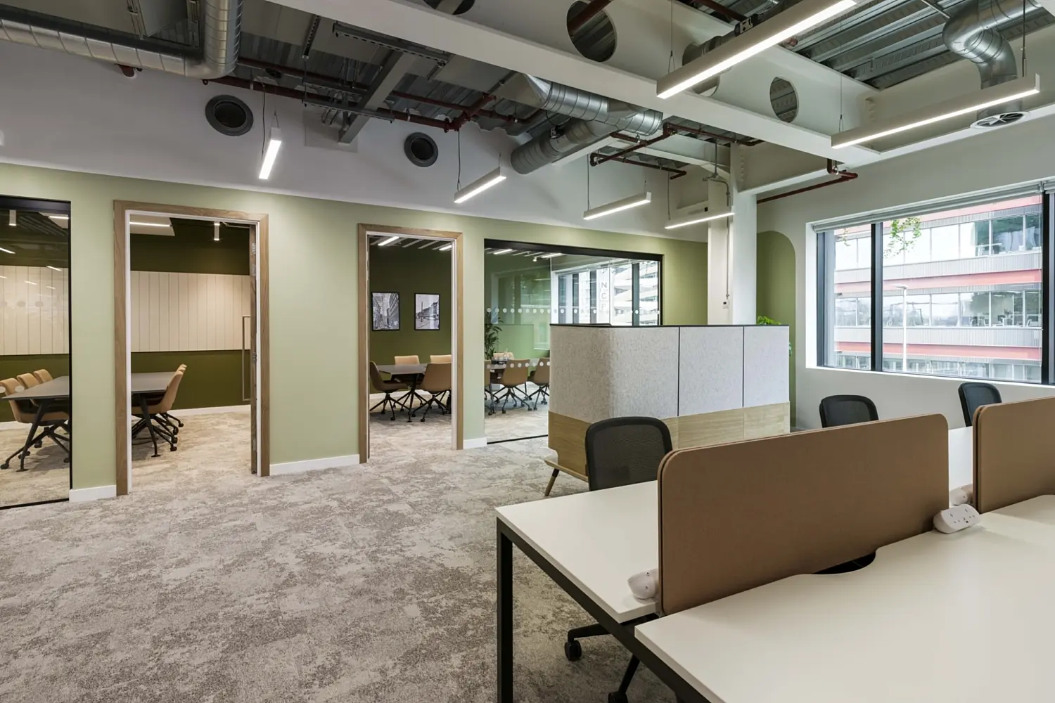 Modern office workspace with a calm, contemporary design and a muted green and neutral colour palette. In the foreground, white desks with soft pink privacy screens and black office chairs are arranged in a cluster on a patterned carpet.  Along the left side, three open doorways lead into enclosed meeting rooms, each fitted with tables and matching chairs, with glass panels allowing visibility into the spaces. Between the meeting rooms and desks, a low storage unit with a speckled finish and wooden base sits centrally, topped with plants.  Large windows on the right bring in natural light and offer views of a neighbouring building. Overhead, the ceiling is exposed, showing ductwork, pipes and suspended linear lighting, reinforcing the industrial aesthetic. The combination of green walls, soft furnishings and planting creates a balanced, modern working environment.