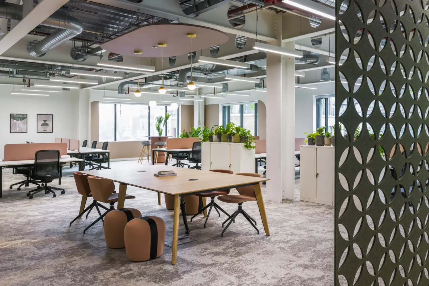 Modern open-plan office space with a soft, contemporary design. In the foreground, a large rectangular wooden table is surrounded by upholstered chairs in warm brown tones, with two small ottoman-style stools positioned beside it. Beyond, rows of desks with office chairs are arranged in clusters, divided by low white storage units topped with green plants.  Large windows along the back wall allow natural light to fill the space, complemented by a mix of linear ceiling lights and three round pendant lights suspended beneath a circular pink ceiling feature. The ceiling is exposed, showing silver ductwork, pipes and structural elements, giving an industrial feel.  On the right side, a decorative partition with repeating circular cut-outs frames the scene and adds texture. The carpeted floor has a subtle patterned finish in neutral tones, reinforcing the calm, modern working environment.