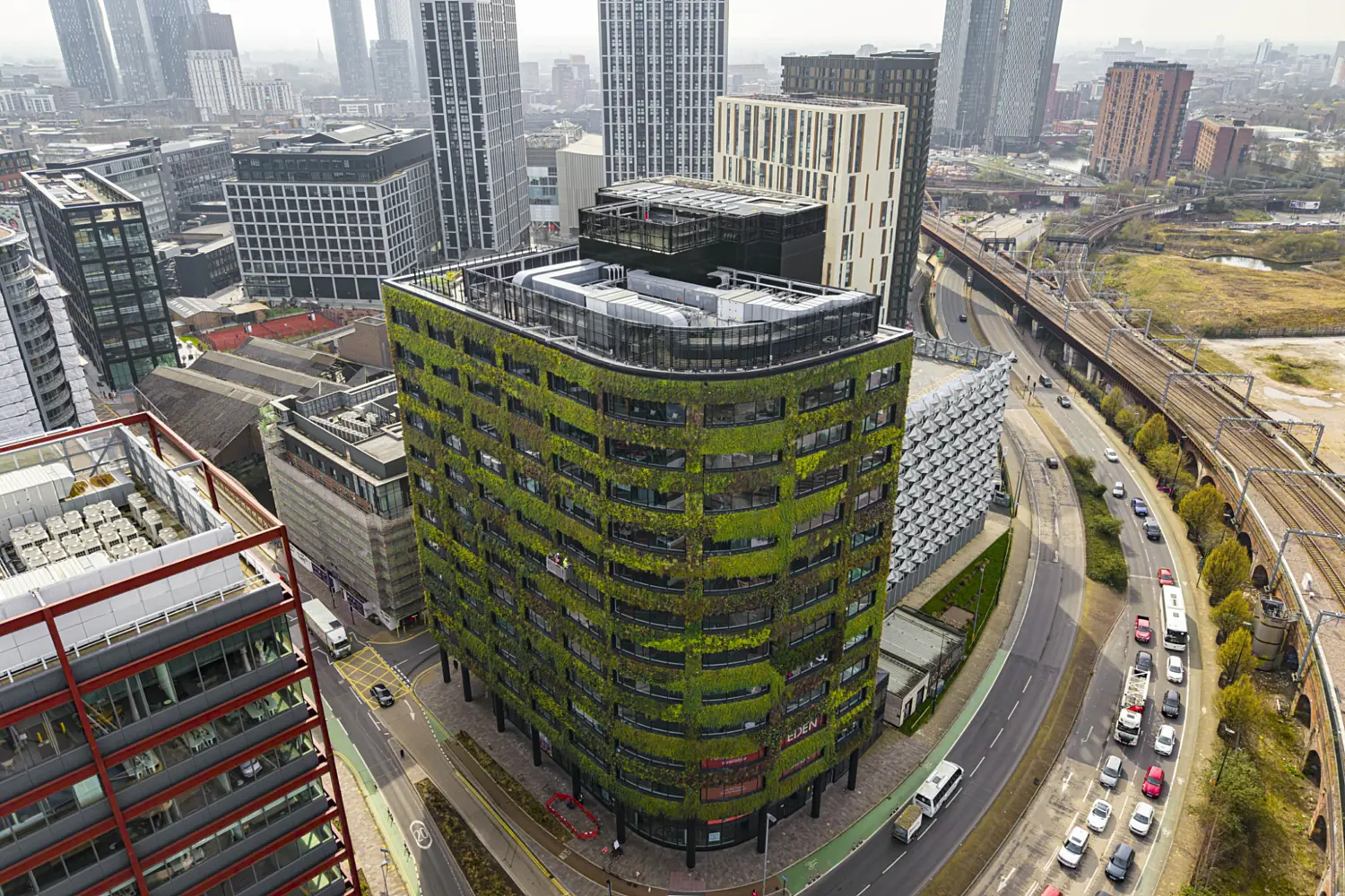Aerial view of a city district featuring a mid rise office building wrapped in green living walls, with plants covering much of the façade. The building sits at a curved road with cars and buses moving along multiple lanes, while railway tracks run parallel on the right. Surrounding the scene are tall modern towers, glass fronted offices and construction areas. The sky appears hazy, and rooftops show plant equipment and terraces on several buildings.