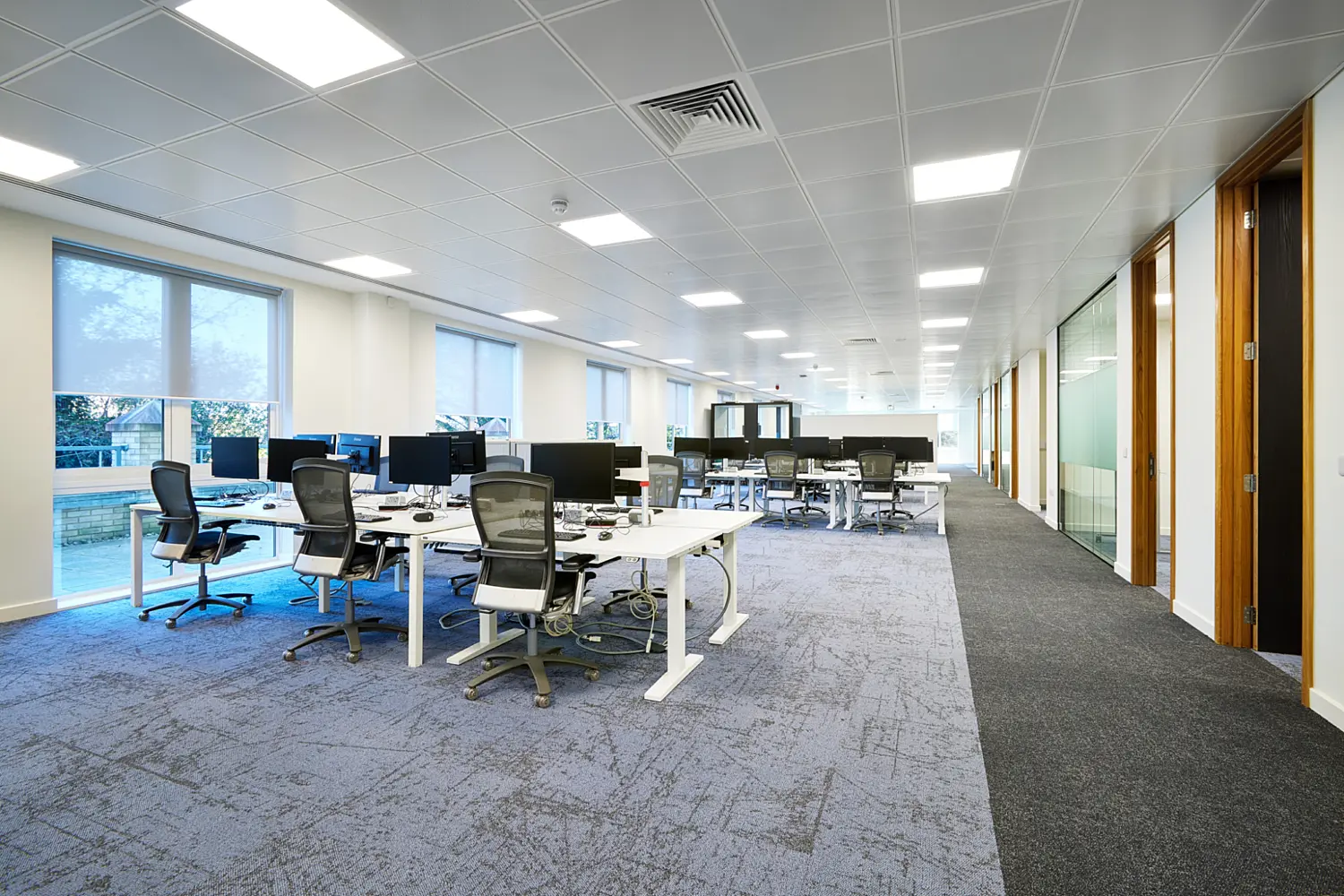 Open-plan office workspace with clusters of white desks, computer monitors and ergonomic mesh chairs arranged across a large carpeted floor. The bright interior features floor-to-ceiling windows, recessed ceiling lighting and glazed meeting rooms with timber-framed doorways along the right-hand side.