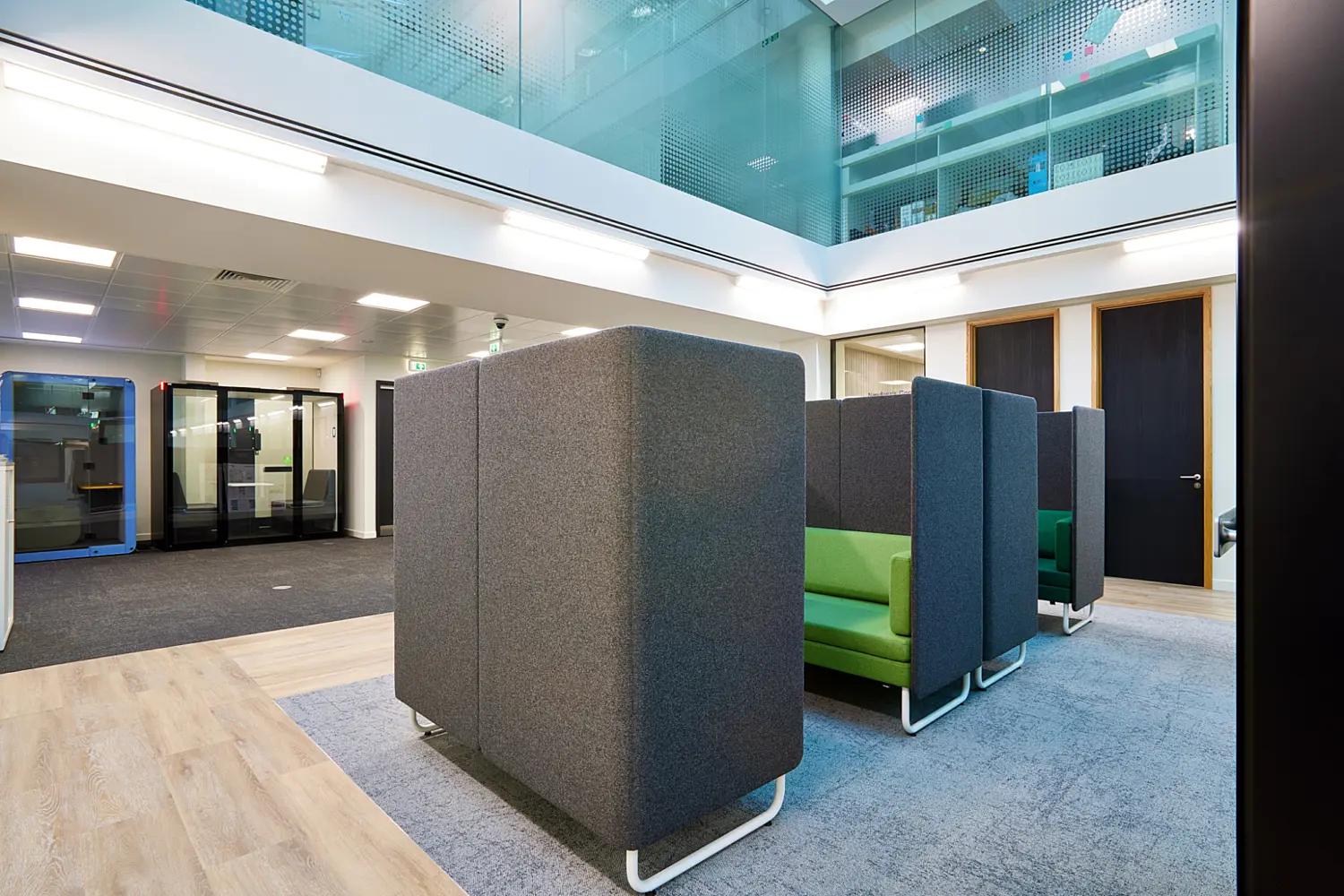 Open-plan office breakout area with high-backed acoustic seating pods in grey fabric and green upholstery, set beneath a glass atrium. The space features carpet and wood-effect flooring, private meeting booths in the background and modern office doors along the right-hand wall.