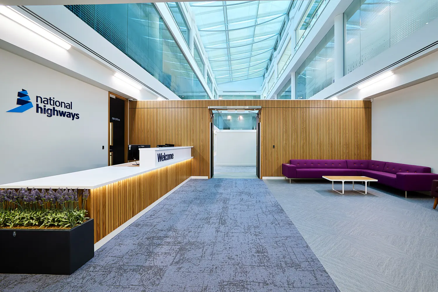 Modern office reception area with a white welcome desk, timber slatted wall panels and a large glass atrium overhead. The National Highways logo is displayed on the left wall, while purple modular seating and a low table sit on the right side of the lobby.