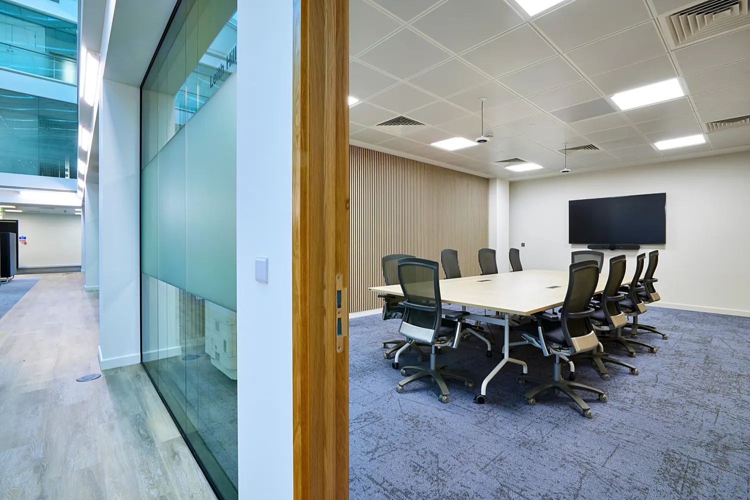 Modern meeting room with a light wood conference table surrounded by black mesh office chairs, viewed through an open doorway beside a glazed corridor. The room features grey carpet flooring, timber slatted wall panelling, recessed ceiling lights and a large wall-mounted display screen.