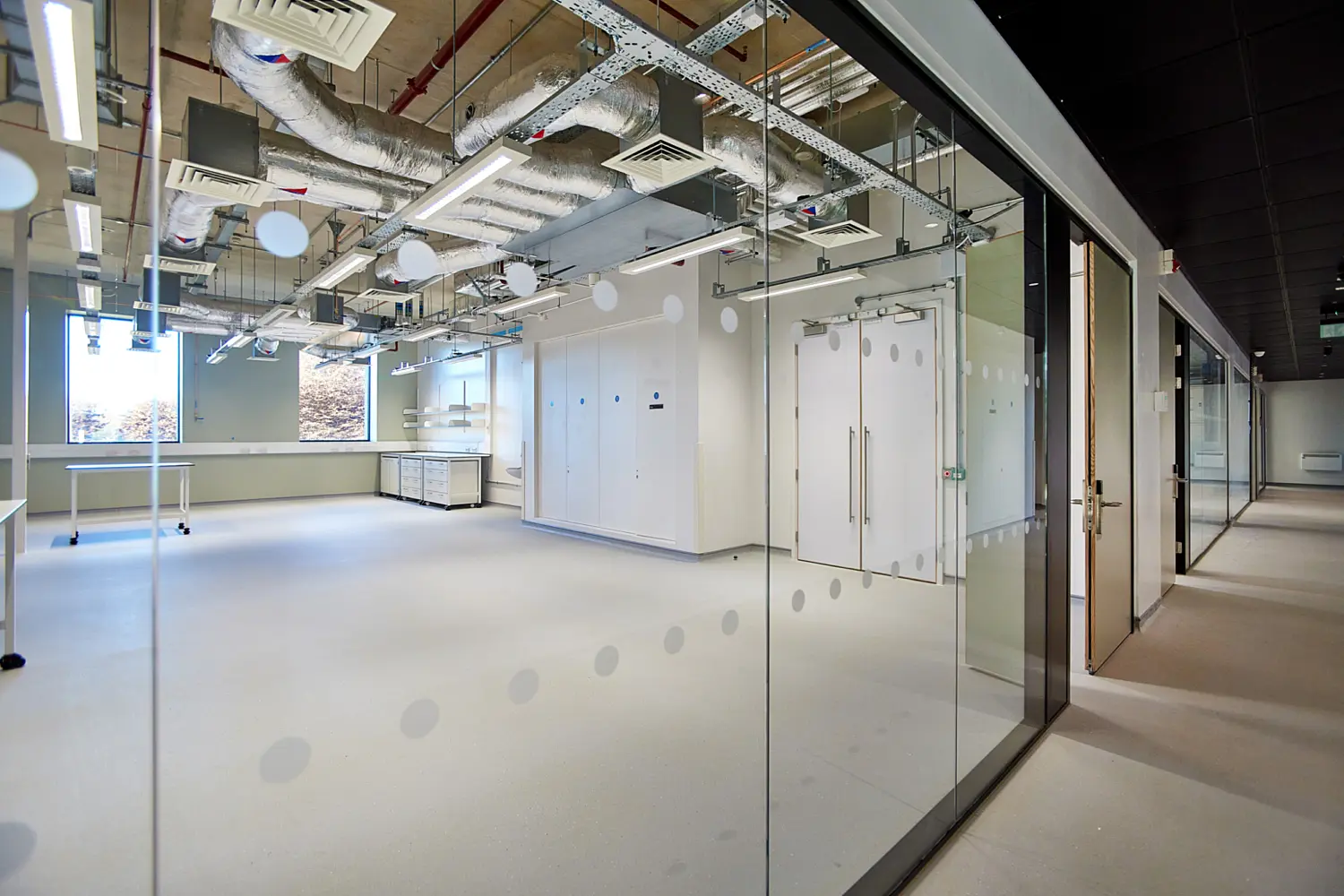 View into a modern laboratory through a glass partition, showing white floors, built-in storage cabinets, mobile worktables and extensive exposed ceiling services, with a corridor running alongside the glazed wall.