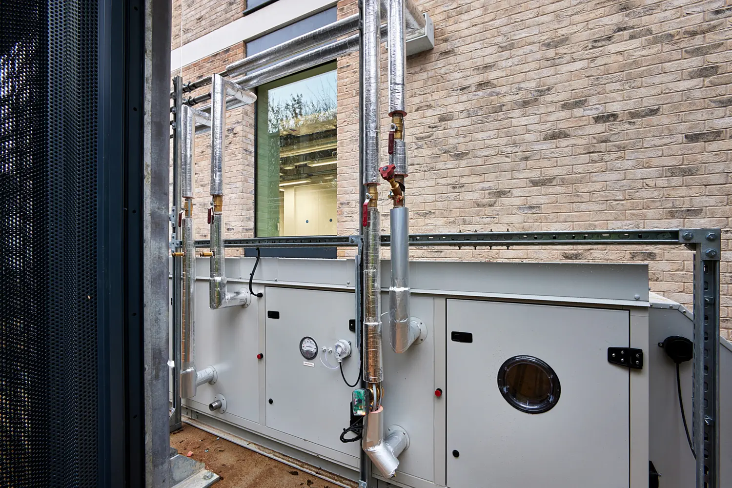 Exterior mechanical equipment installation beside a brick building, showing a large grey air handling unit with connected metal ducting and insulated pipework, mounted within a steel frame near a window.