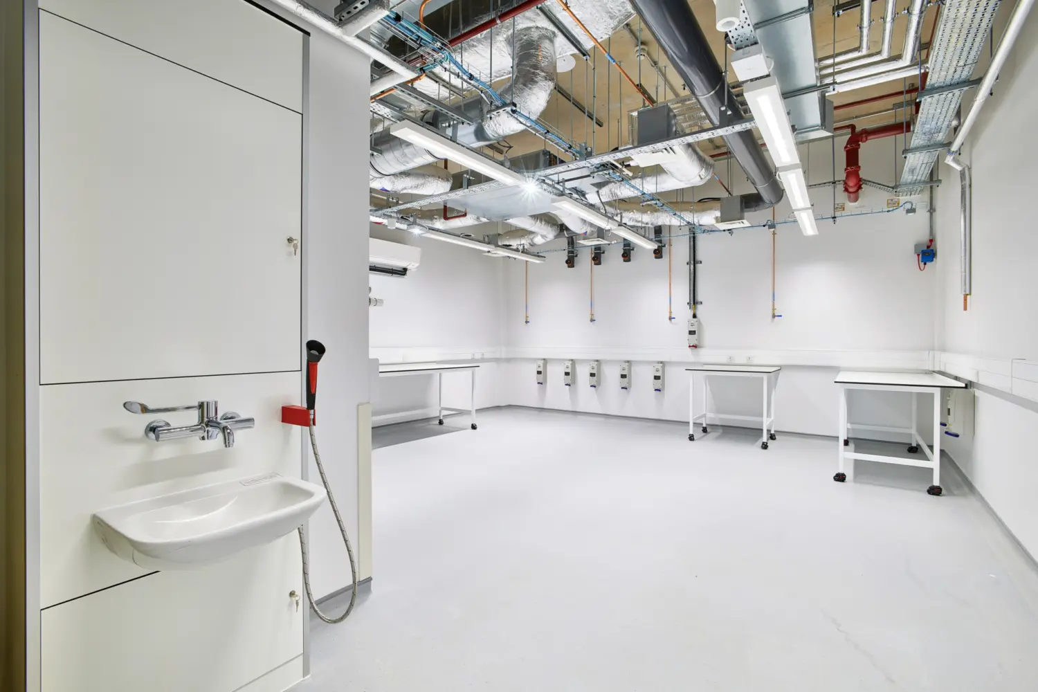 A bright, empty laboratory space with white walls and flooring, mobile white workbenches around the room, a wall-mounted sink with hand spray in the foreground and exposed ducts, pipes and lighting visible in the ceiling.