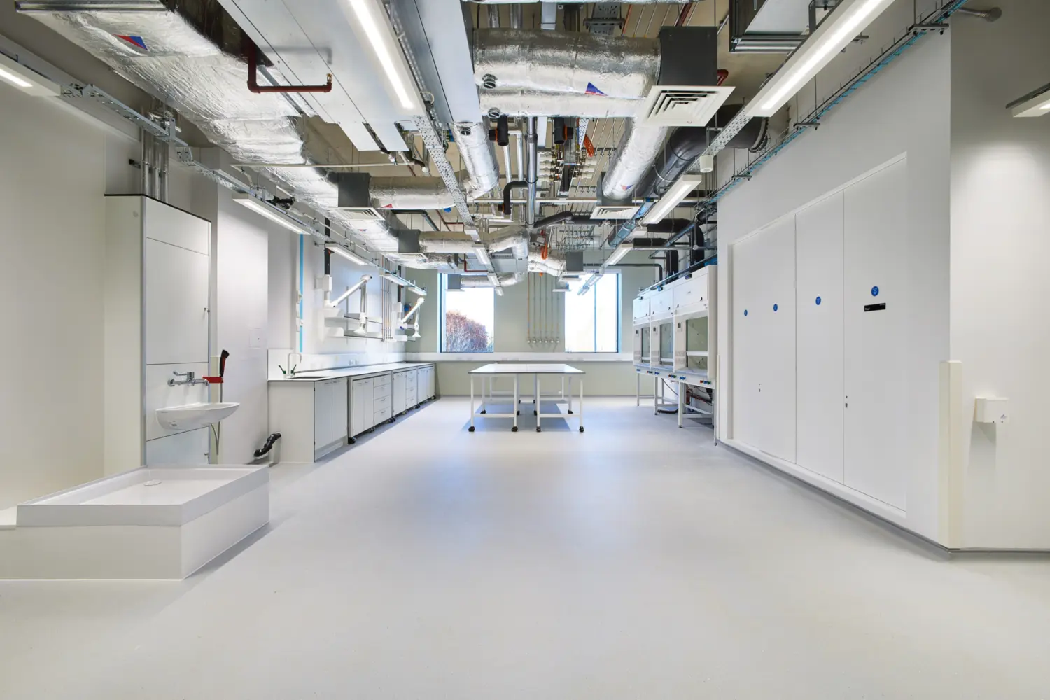 Bright, empty laboratory interior with white floors and cabinets, a central worktable and exposed ceiling services including ducts, pipes and lighting, facing a window at the far end.