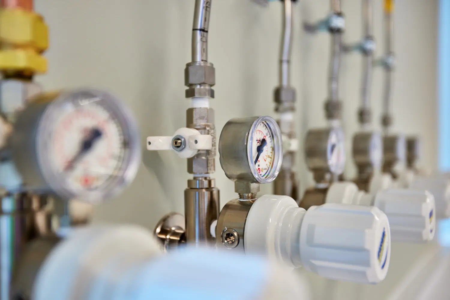 Close-up of industrial pipework with metal valves, pressure gauges and white control knobs mounted in a row against a light-coloured wall, showing a technical services installation in sharp focus.