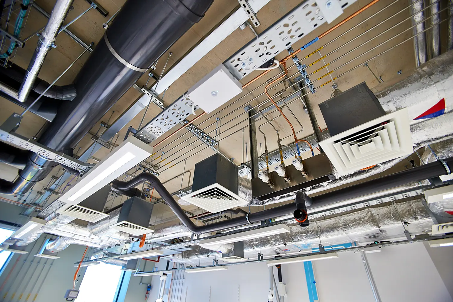 Exposed laboratory ceiling showing ventilation ductwork, cable trays, pipes, lighting fixtures and air diffusers mounted beneath a concrete soffit as part of the building services installation.