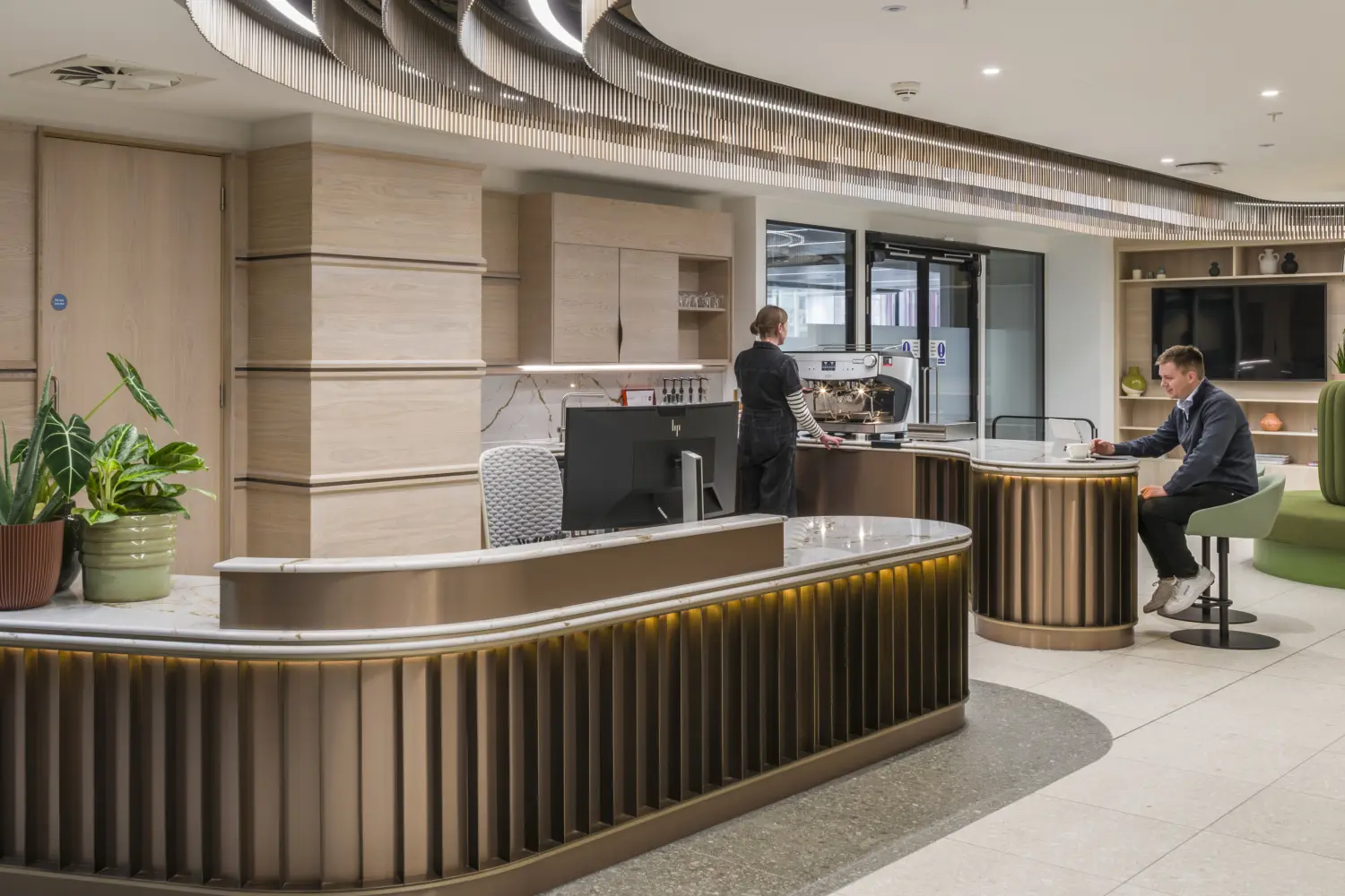 Contemporary office reception and hospitality area featuring a curved reception desk with illuminated bronze-toned detailing and a matching coffee bar. Light wood cabinetry, stone surfaces and integrated shelving create a refined, hotel-inspired aesthetic. A barista-style coffee machine sits at the refreshment counter, while casual seating and planting are arranged throughout the space. Overhead, a sculptural ceiling feature with curved metallic fins and integrated lighting mirrors the flowing forms of the furniture below, creating a welcoming environment for visitors, meetings and informal work.