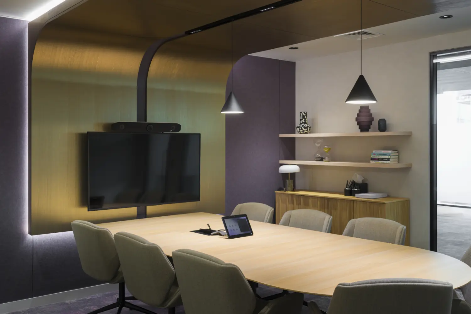 Contemporary meeting room with a large oval timber conference table surrounded by upholstered chairs. A wall-mounted display screen with integrated video conferencing equipment is positioned against a feature wall finished with metallic panels and recessed lighting. Decorative shelving, pendant lights and timber storage units add a hospitality-inspired feel, while acoustic wall finishes and soft carpeting create a comfortable setting for formal meetings and hybrid collaboration.