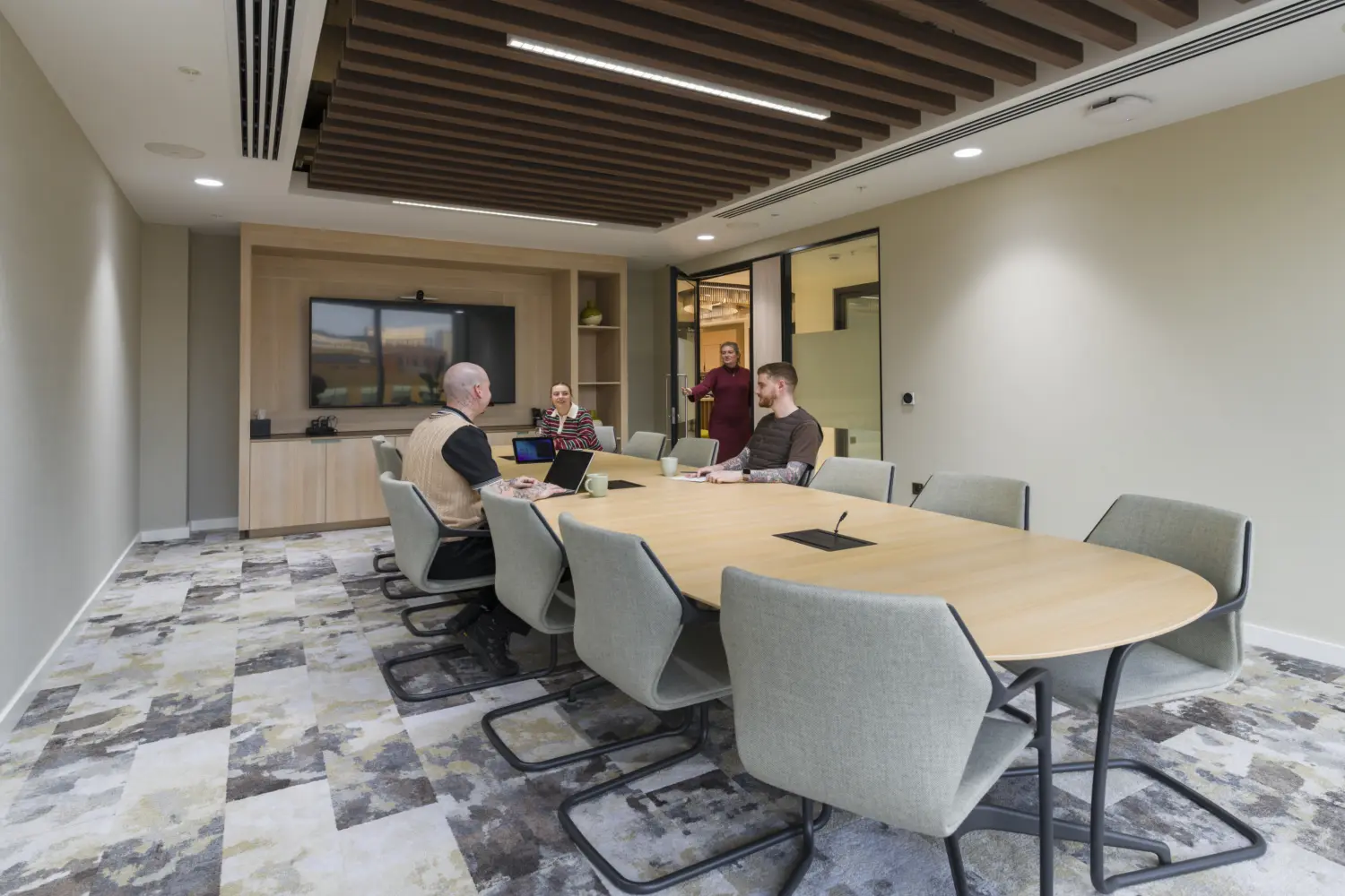 Modern boardroom with a large oval timber meeting table surrounded by upholstered conference chairs. A wall-mounted display screen with integrated video conferencing equipment is positioned within bespoke light wood joinery at one end of the room. Several people are seated around the table using laptops, while another person stands in the doorway. The space features a patterned carpet, acoustic timber ceiling slats, neutral wall finishes and glazed partitions, creating a professional environment for meetings and collaboration.