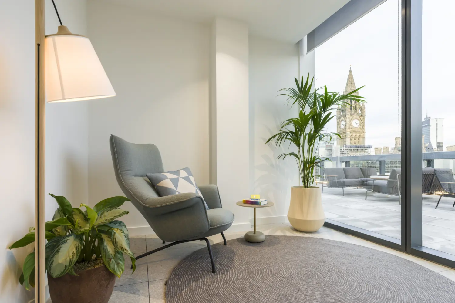 Quiet office lounge area with a high-backed upholstered armchair, floor lamp and side table arranged on a circular rug beside floor-to-ceiling glazing. Several indoor plants add greenery to the space, while an outdoor rooftop terrace with seating is visible through the windows. A historic clock tower can be seen in the background beyond the terrace. The space provides a comfortable setting for informal work, reading and relaxation, with abundant natural light and panoramic city views.