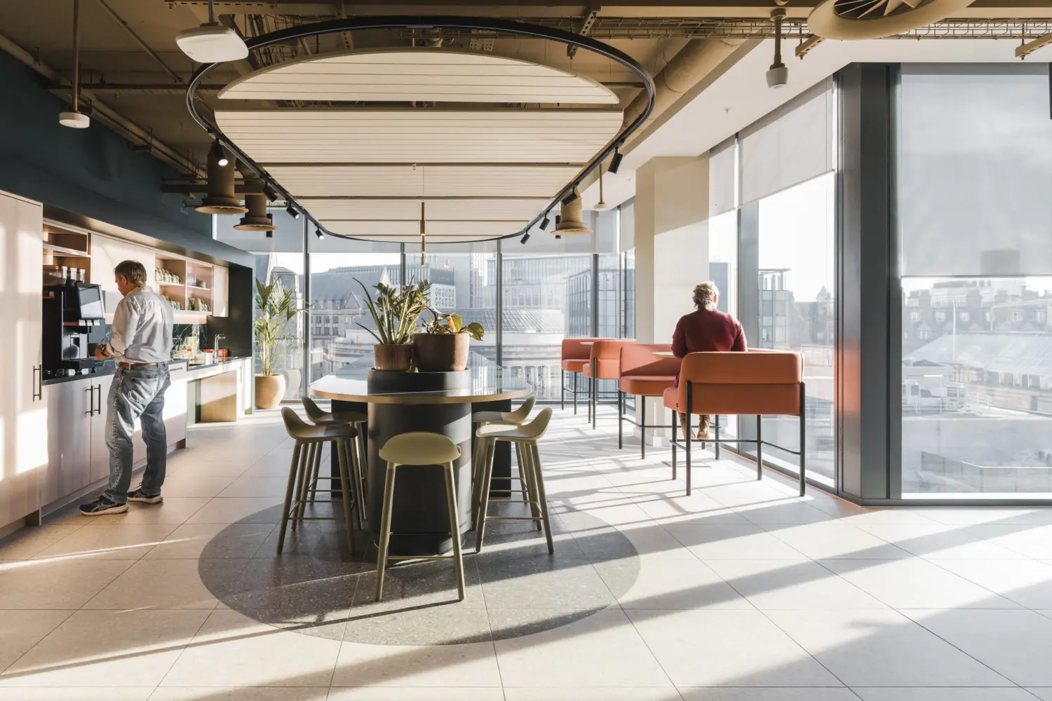 Modern office café and breakout space with floor-to-ceiling windows offering views across the city. A central round table with integrated planting is surrounded by stools, while high-backed booth seating lines the window perimeter. A refreshment area with coffee machines, shelving and storage occupies one side of the room. The interior features large ceiling-mounted acoustic panels, exposed services, light tiled flooring and abundant natural daylight, creating a bright and informal setting for breaks, collaboration and social interaction.