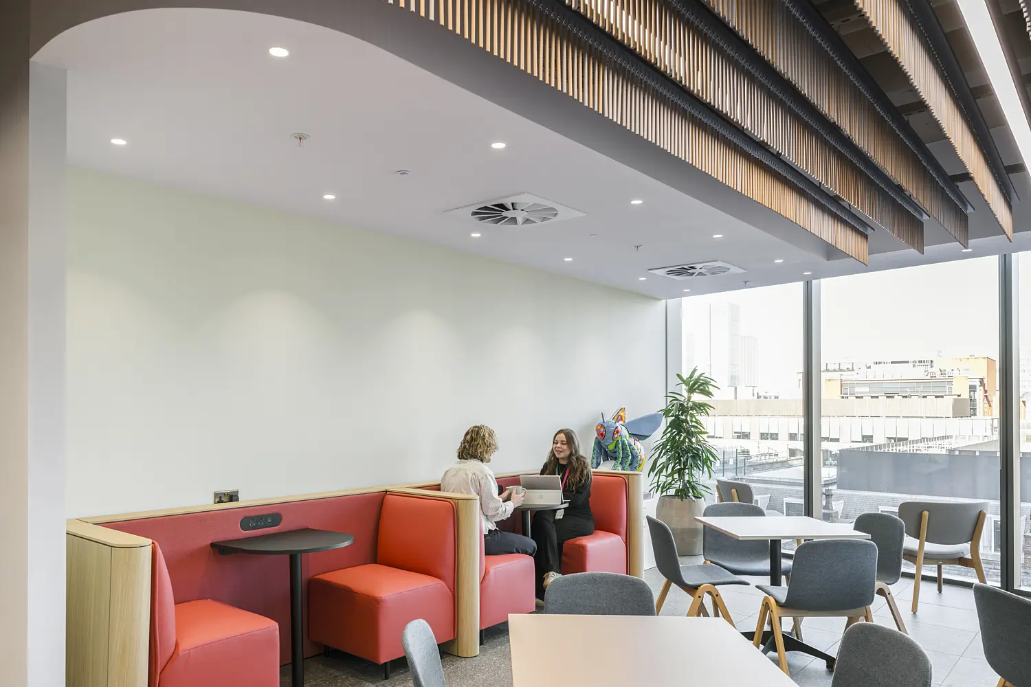 Contemporary office café and collaboration area featuring red upholstered booth seating, freestanding tables and grey upholstered chairs arranged beside floor-to-ceiling windows. Two people are seated in one of the booths using a laptop, while additional café-style seating is positioned throughout the space. A striking ceiling feature with curved bronze-toned fins and integrated lighting runs overhead, creating a strong architectural focal point. Natural daylight fills the area, which combines hospitality-inspired furnishings, city views and flexible settings for informal meetings, dining and focused work.