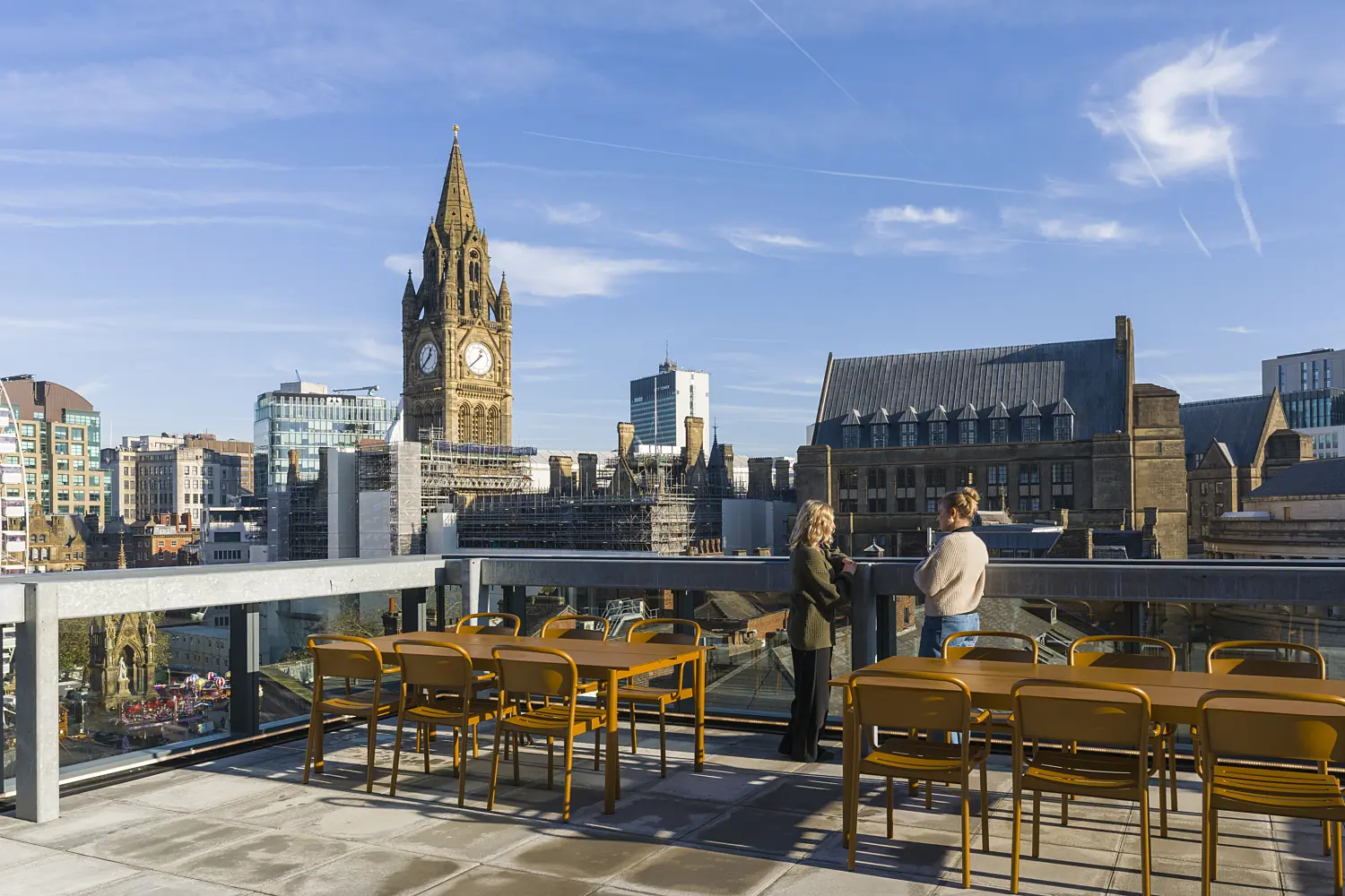 Rooftop terrace with outdoor dining tables and chairs overlooking a city skyline. Two people stand beside a glass balustrade, with a historic clock tower and surrounding stone buildings forming a prominent backdrop. A large observation wheel is visible in the distance, while clear blue skies and bright daylight illuminate the space. The terrace provides an outdoor setting for informal meetings, breaks and social gatherings with panoramic urban views.