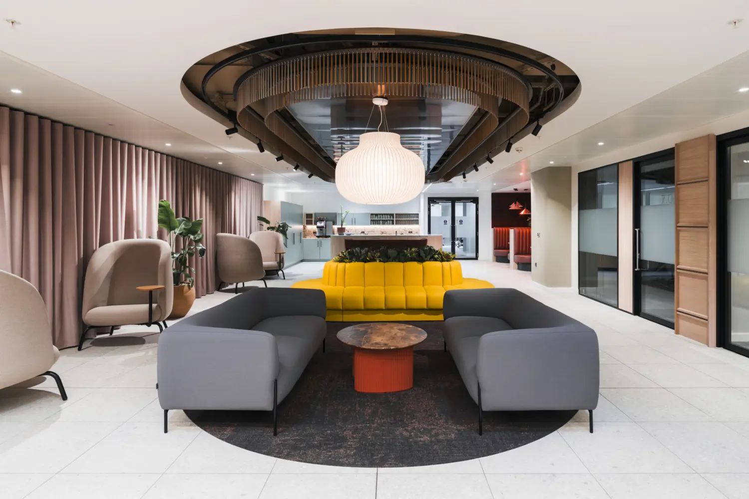 Contemporary office lounge and reception area featuring curved grey sofas arranged around a circular coffee table, with a bright yellow banquette and integrated planting at the centre. A large decorative pendant light hangs beneath a sculptural ceiling feature with timber detailing and black accents. Soft lounge chairs line the perimeter beside full-height curtains, while glazed meeting rooms and a refreshment area are visible in the background. The space combines light stone flooring, warm neutral tones and modern furnishings to create a welcoming hospitality-inspired workplace environment.