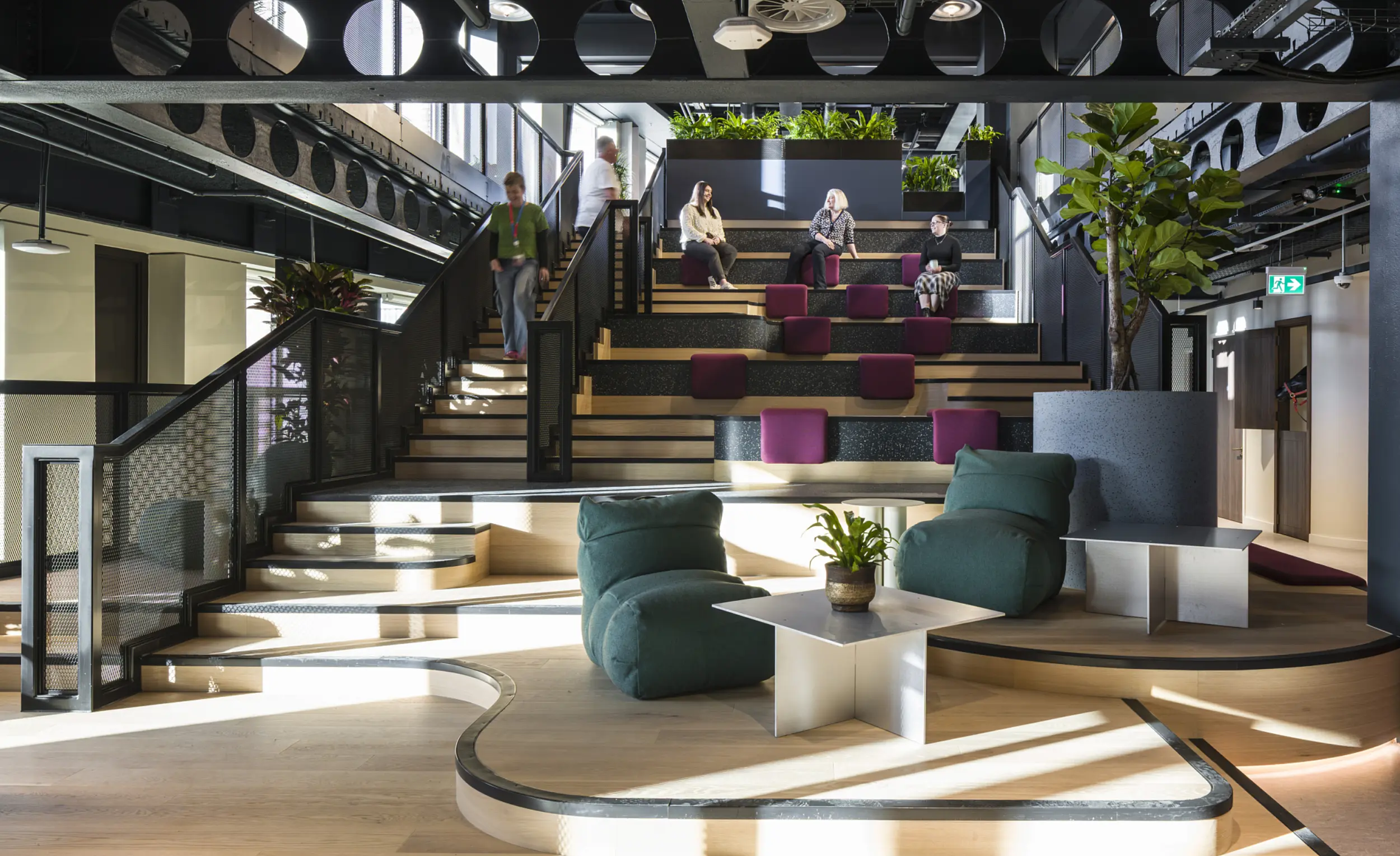A multi level workplace space featuring tiered seating with purple cushions, surrounded by planting and framed by black steel beams. Curved wooden platforms with low lounge chairs and small white tables create informal seating on the lower level. Several people sit and walk through the space. Large planters and full height windows bring in greenery and natural light.