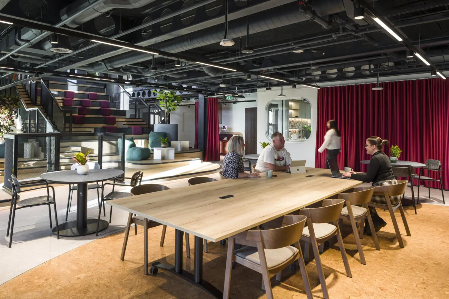 A social workspace with a large wooden table and simple chairs in the foreground where several people sit and talk. To the left, round café tables sit near tiered seating with colourful cushions. A red curtain spans the back wall and the ceiling features exposed services with linear lighting. Planting and soft seating are arranged throughout the space, creating a relaxed, open environment.