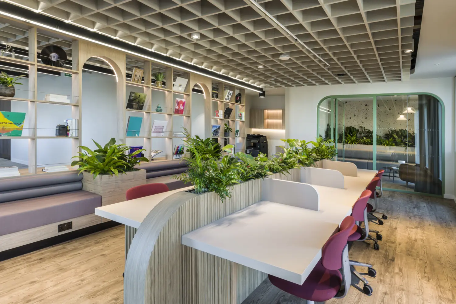 A bright workspace with a long white desk and red task chairs separated by low dividers topped with plants. To the left, built in seating sits below open shelving with arched frames displaying books and objects. A gridded ceiling feature runs across the space. At the back, a corridor with curved glass doors leads to an area filled with more planting.