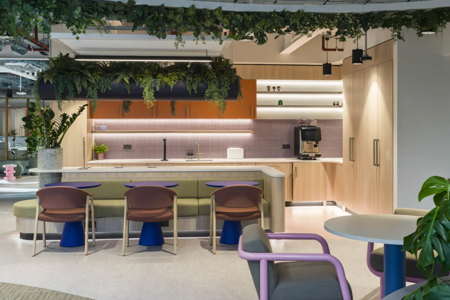 A workplace tea point and social area featuring a central island with a white countertop and soft seating in front. Three upholstered stools with blue bases sit along the island. Above, a suspended planter filled with dense greenery hangs over the counter. The back wall includes timber cabinets, open shelving and a tiled splashback with integrated lighting. To the right, lounge chairs and a small round table create a relaxed seating spot among more planting.
