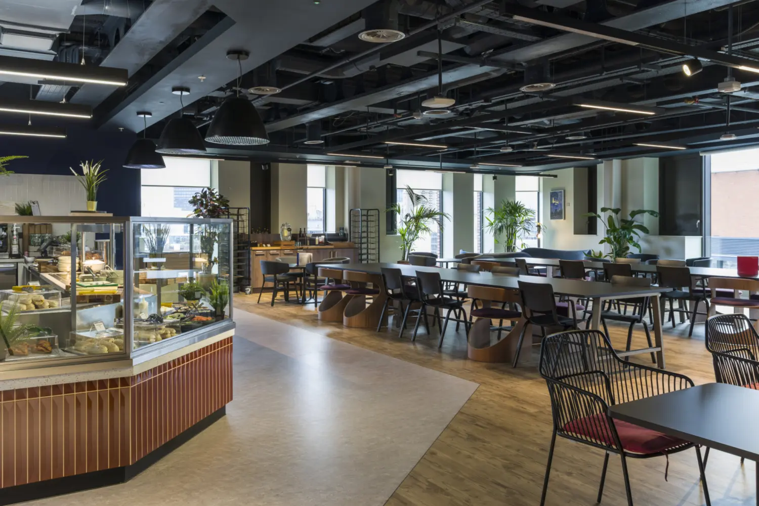 A workplace dining area with a central food counter and glass display filled with dishes on the left. The space includes long wooden tables with dark chairs arranged across a floor that mixes wood and light vinyl. Large pendant lights hang from an exposed ceiling. Tall plants are placed throughout, and full height windows bring daylight into the room.