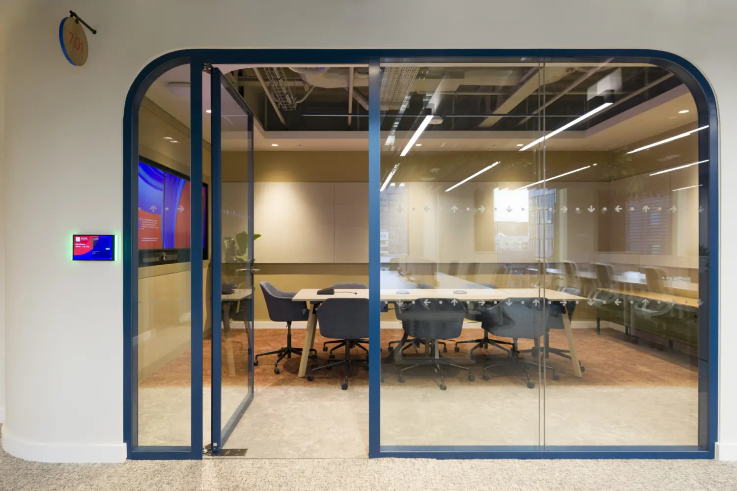 A glass fronted meeting room with rounded blue framing, showing a long table with upholstered chairs arranged around it. Two large screens are mounted on the far wall. The ceiling features exposed services and linear lighting. A room booking panel is mounted on the wall outside, and the glass panels reveal parts of the surrounding workspace.