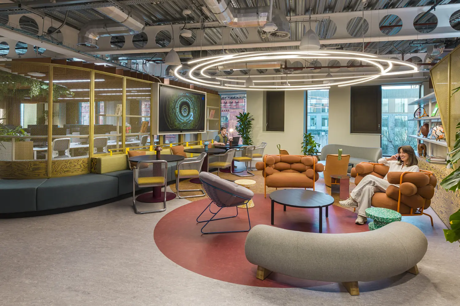 A vibrant open lounge area with mixed seating including curved sofas, round chairs and modular orange seating around circular tables. A large circular LED light feature hangs from an exposed ceiling above a red floor accent. To the left, a glazed meeting space with yellow framing sits behind a curved bench. Planting is placed throughout the space and large windows bring in natural light.