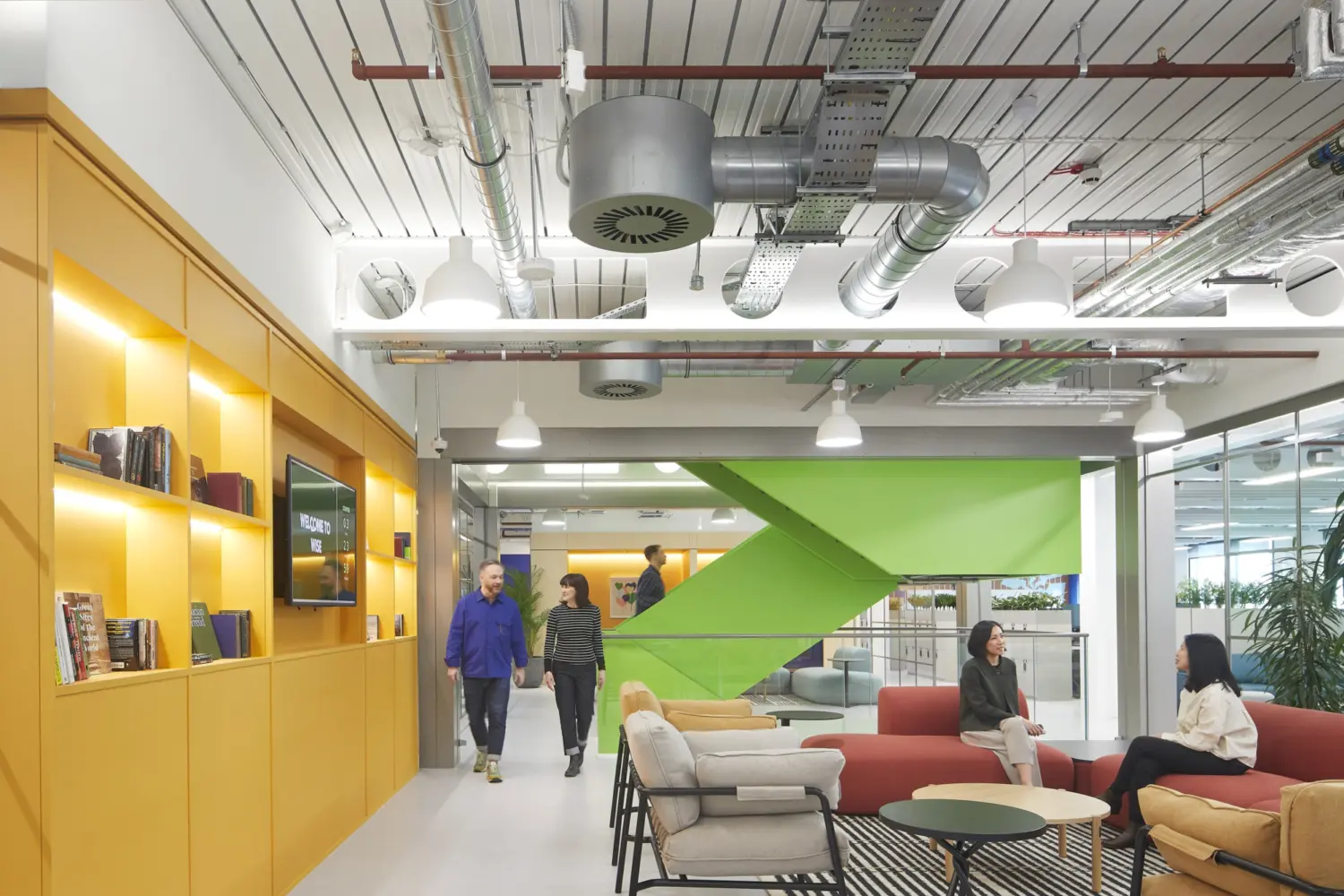 Modern open plan office lounge with exposed ducts and white ceiling, featuring a yellow shelving wall with books and a screen on the left, a green angular feature wall in the centre, and sofas and armchairs arranged around low tables on a patterned rug, with several people sitting and walking through the space.