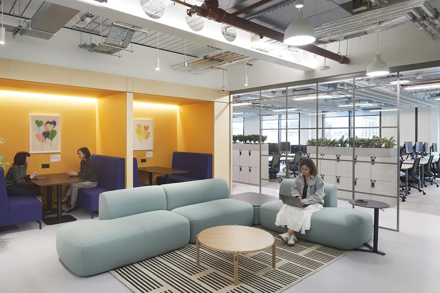 Open plan office seating area with a curved light blue modular sofa and round wooden coffee table on a patterned rug, yellow booth seating built into the wall on the left with two people working at a table, and a glass partition on the right revealing a workstation area with desks, chairs and computers, under a ceiling with exposed services and pendant lighting.