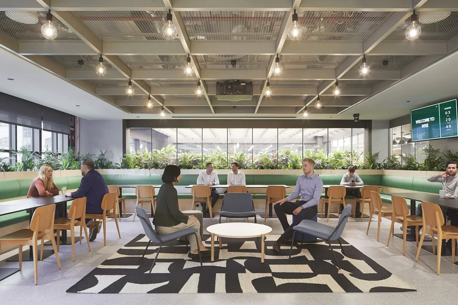 Large open plan office breakout and café style area with long tables and wooden chairs arranged along green upholstered bench seating, a central seating area with armchairs and low tables on a bold patterned rug, abundant planting along the perimeter, and a ceiling with a grid structure and exposed lighting, with multiple people seated and talking throughout the space.